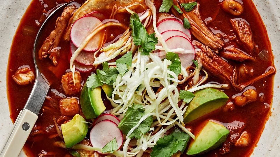 a bowl of homemade turkey pozole rojo topped with seasoned cabbage, avocado, cilantro, radishes, and lime juice.