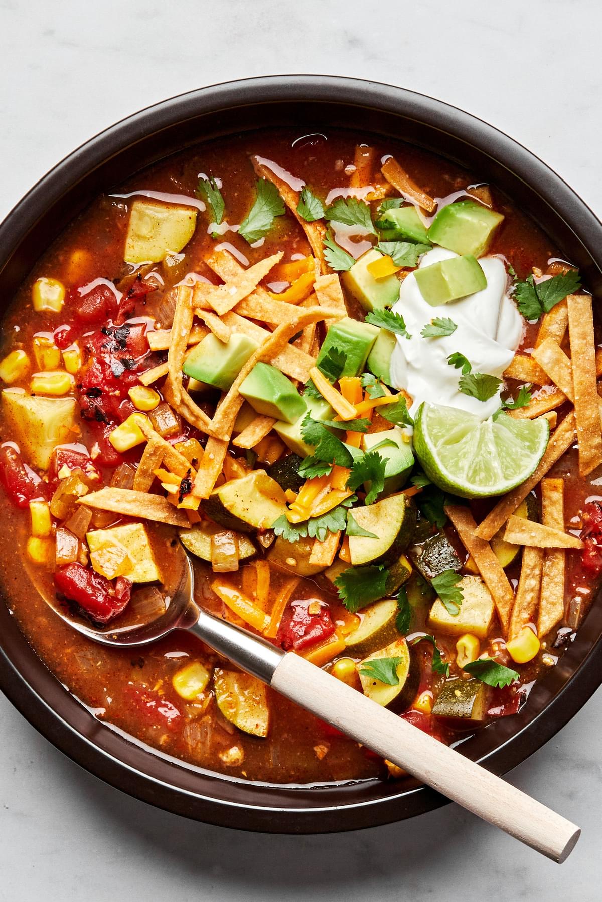 a bowl of vegetarian tortilla soup topped with cheese, sour cream, cilantro, avocado and lime wedges