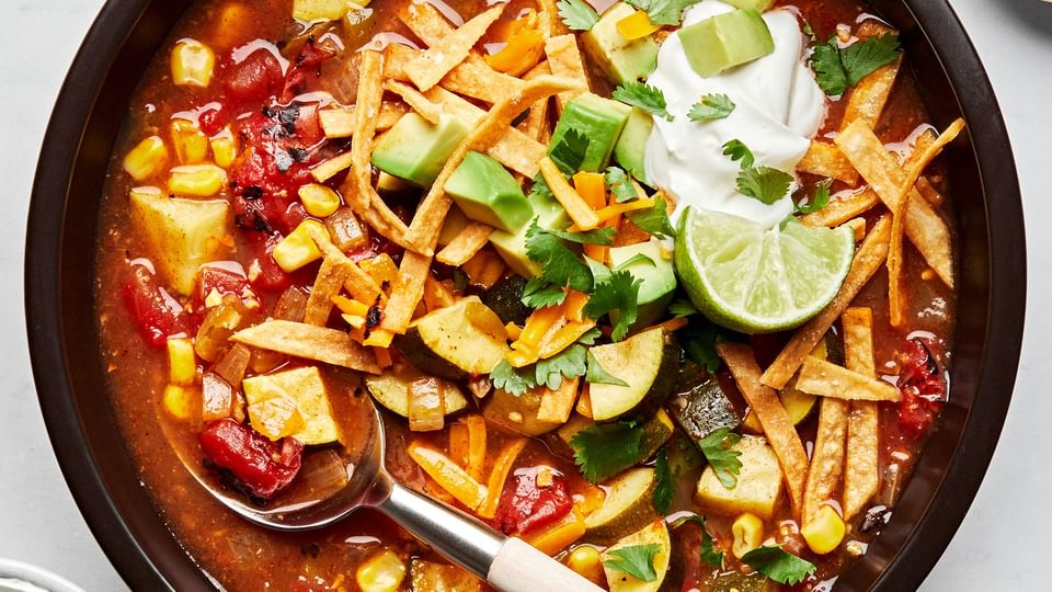 a bowl of vegetarian tortilla soup topped with cheese, sour cream, cilantro, avocado and lime wedges
