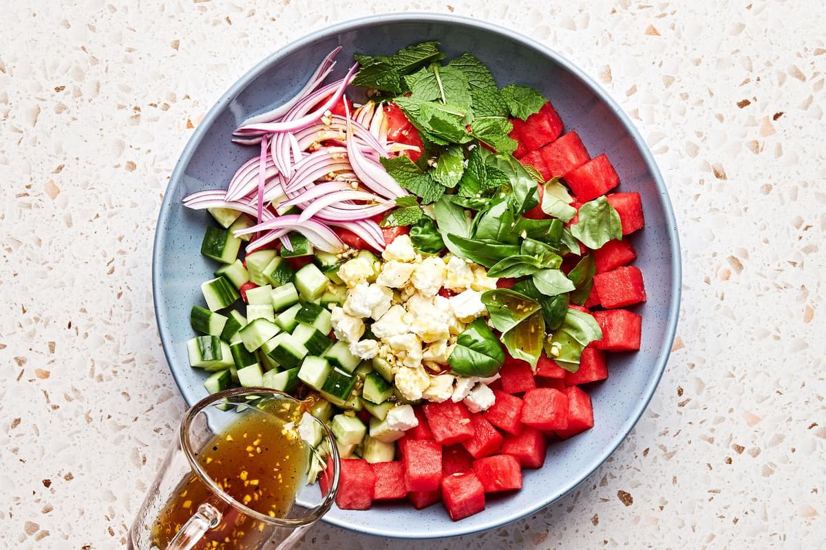 white balsamic dressing being poured over watermelon, cucumber, onions, feta, mint and basil in a bowl