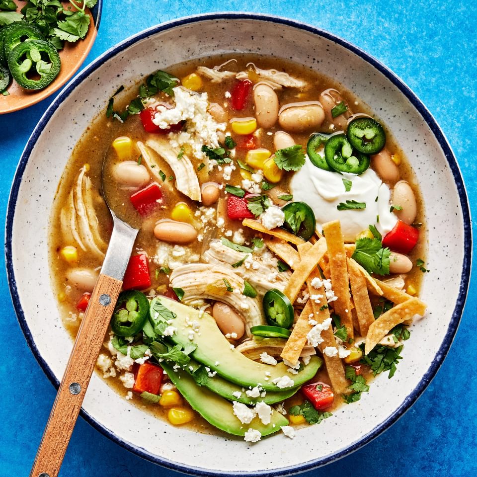 a bowl of white bean chicken chili topped with jalapeño, Sour cream, avocado, tortilla strips, cotija and cilantro