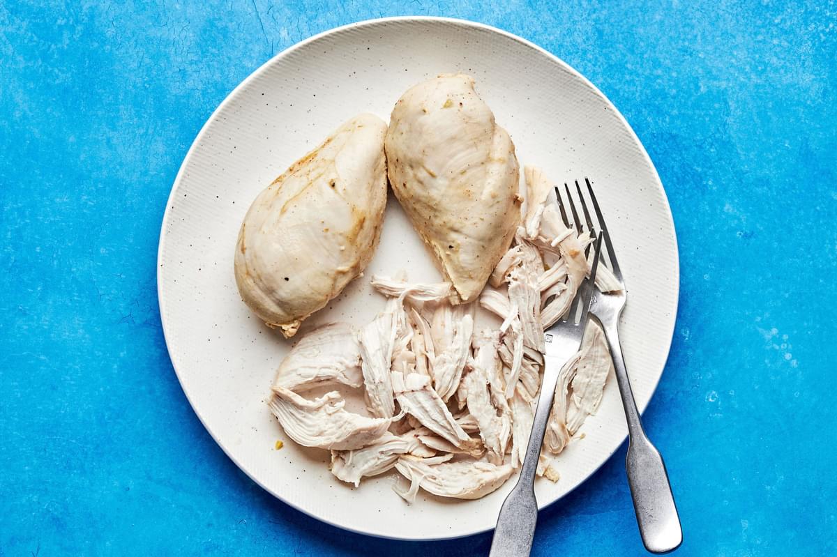 cooked chicken breast being shredded with 2 forks on a plate