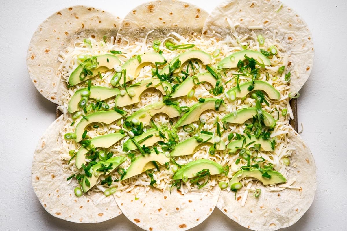flour tortillas on a baking sheet with shredded cheese, avocado and green onions.