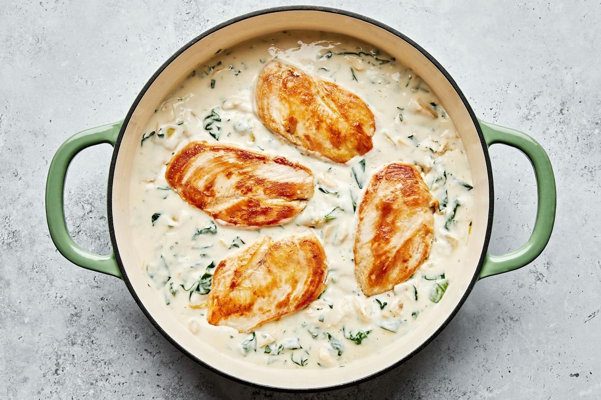 chicken being added to a skillet with spinach artichoke cream sauce