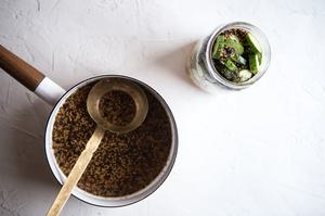 A pot of pickling spices and brine with vinegar and ladle next to jar of sliced cucumbers