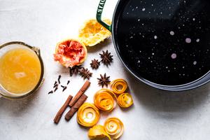 Red wine in large pot next to fresh orange juice, orange peels, cinnamon sticks, whole star anise and whole cloves.