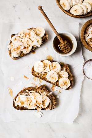 Almond Butter Toast with Bananas, Toasted Coconut, honey and salt on parchment paper