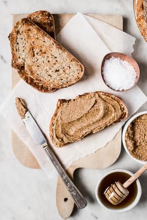 A slice of toasted bread spread with almond butter. Next to a bowl of honey, flakey salt and a knife.