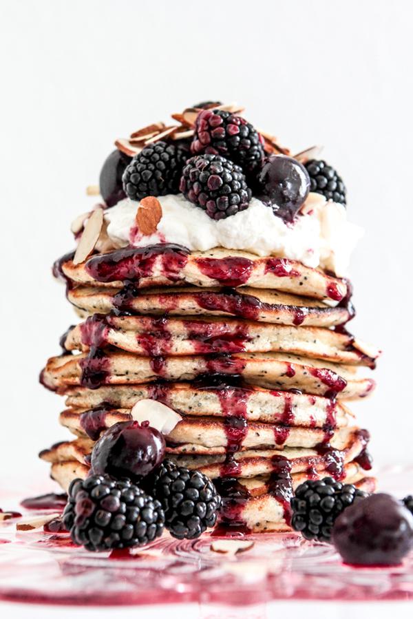 a stack of almond poppy seed pancakes topped with whipped cream, fresh berries and cheery syrup