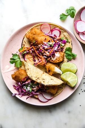 Beer-Battered Fish Tacos with sesame slaw and a spicy cream on a plate with radishes and cilantro