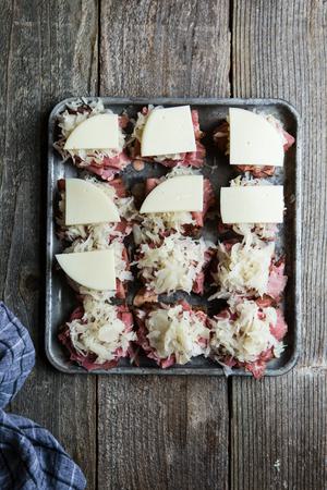 small pieces of rye bread on a baking sheet topped with Russian dressing, pastrami, sauerkraut and sliced provolone cheese