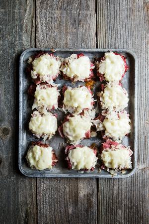 small pieces of rye bread on a baking sheet topped with Russian dressing, pastrami, sauerkraut and melted provolone cheese