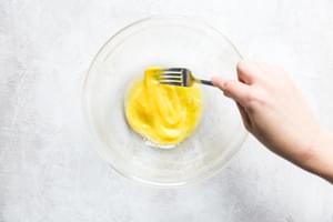 a hand holding a fork whisking 2 eggs in a glass bowl
