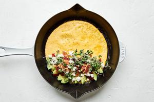an omelette being filled with avocado, goat cheese and bacon cooking in a skillet