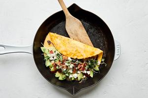 an omelette filled with avocado, goat cheese and bacon being folded in half in a skillet