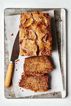a loaf of homemade banana bread topped with banana slices and granulated sugar being sliced with a knife