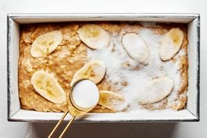 banana bread dough in a prepared loaf pan being topped with thinly sliced bananas and sprinkled with granulated sugar