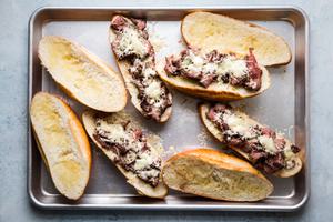 homemade french dip sandwiches on a baking sheet with thinly sliced steak, garlic butter and melted gruyere cheese