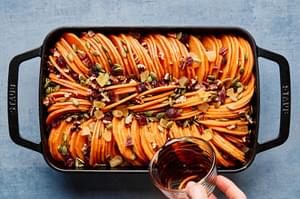 maple syrup being drizzled over seasoned sweet potatoes in a baking dish