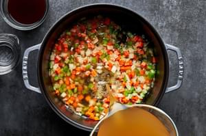 chicken broth being poured into a pot with onion, carrots, celery, and salt