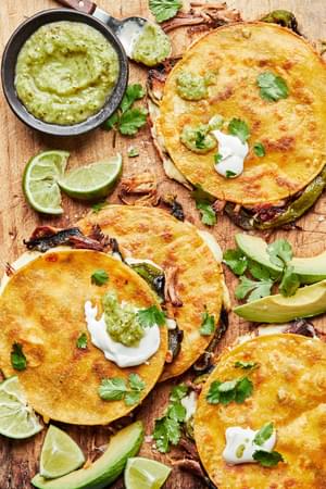Roasted Poblano and Carnitas Quesadillas on a cutting board surrounded by lime wedges, salsa verde and cilantro
