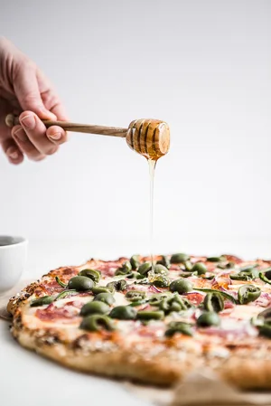 honey being poured on Salami, Jalapeño & Olive Pizza with , jalapeños, red onions and everything bagel crust