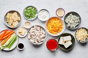 ingredients to make buffalo chicken dip in bowls on the counter including chicken, cheese, spices and buffalo sauce