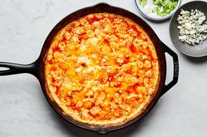 homemade buffalo chicken dip in a cast iron skillet fresh out of the oven next to bowls of green onions and blue cheese