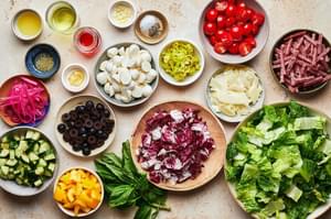 Ingredients for homemade Italian chopped salad in bowls on the counter including lettuce, salami, veggies, and mozzarella