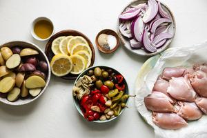 raw chicken on a plate, next to raw potatoes, raw onions, lemons, olives and seasoning.
