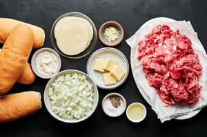 hoagie rolls, sliced ribeye, onions, butter, salt, garlic, pepper, mayo and provolone in bowls to make philly cheesesteaks