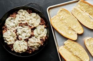cooked beef topped with melted provolone cheese in a cast iron skillet next to a baking sheet with open hoagie rolls