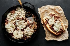 cooked beef topped with melted provolone cheese in a cast iron skillet being scooped onto a hoagie roll on top of parchment