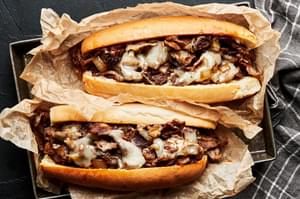 Two homemade Philly cheesesteak sandwiches on hoagie rolls wrapped in parchment paper on a baking sheet