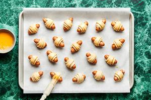 raw pigs in a blanket on a parchment lined baking sheet being brushed with beaten egg