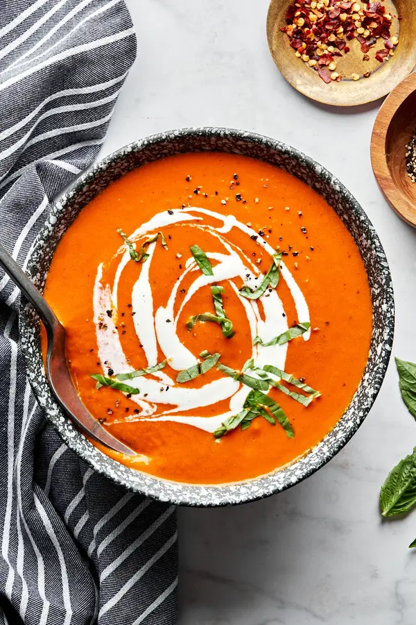 a bowl of homemade tomato bisque with a spoon garnished with a drizzle of cream and fresh chopped basil
