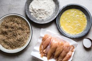 A dish of pecans and breadcrumbs, flour and eggs and chicken tenders on parchment paper
