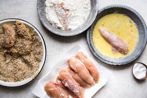 A dish of pecans and breadcrumbs, flour and eggs and chicken tenders on parchment paper with one chicken tender in the eggs