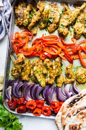 sheet pan chicken shawarma dinner with red onions and red bell peppers next to pita and parsley
