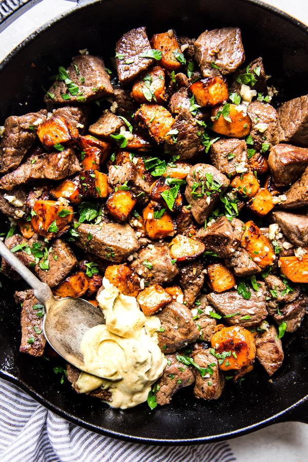 Garlic steak bites with sweet potatoes in a cast iron pan with curry aioli on a spoon.
