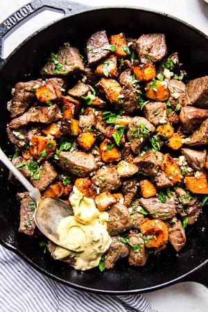 Garlic steak bites recipe with sweet potatoes in a cast iron pan with curry aioli on a spoon.