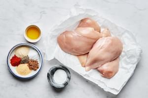 raw chicken on parchment paper next to bowls of olive oil, salt, pepper, garlic powder, onion powder, paprika and brown sugar