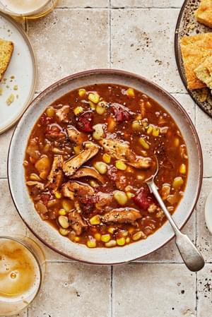 a bowl of homemade Brunswick stew with a spoon next to a small bowl of cracked pepper for serving and a plate of cornbread