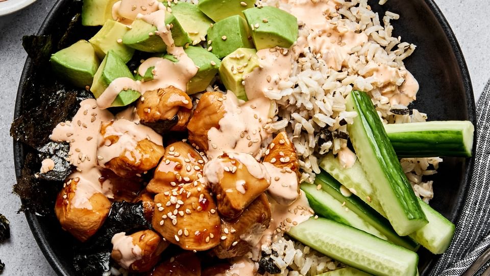 Teriyaki chicken sushi bowl with sriracha mayo, cucumber, avocado and sesame seeds on a bed of seasoned brown rice