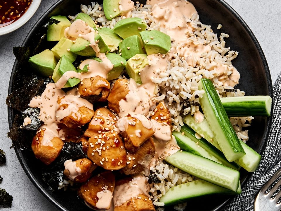 Teriyaki chicken sushi bowl with sriracha mayo, cucumber, avocado and sesame seeds on a bed of seasoned brown rice