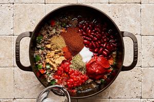kidney beans, canned tomatoes, tomato paste, and spices being added to a pot with cooked ground beef, onions & peppers