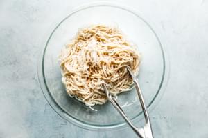 glass bowl with rice noodles and thai peanut dressing being tossed together with cooking tongs