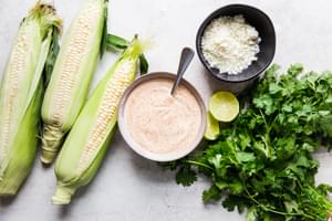 Ingredients for Elote (Mexican Street Corn) laid out: corn on the cob, cilantro, cotija, chipotle mayo and limes