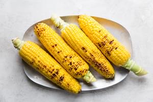 Four grilled ears of corn on the cob on a plate, lightly charred and ready to be prepared as Elote (Mexican street corn).