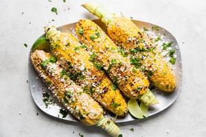 Elote (Mexican street corn) on a platter, topped with chipotle mayo, cotija cheese, and cilantro, served with lime wedges.